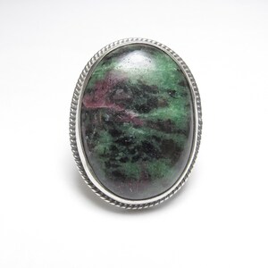 May include: An oval ring featuring a polished cabochon stone with a mottled pattern of deep green, black, and magenta inclusions. The stone is set within a silver-toned metal bezel that has a decorative twisted rope texture along the outer edge.