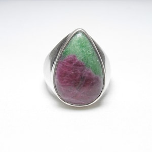 May include: A silver ring featuring a large, teardrop-shaped stone. The stone is a mix of deep red and green hues, creating a striking contrast. The ring has a simple, polished band.