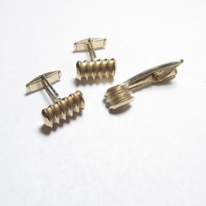 May include: A set of gold-toned cufflinks and a tie clip. The cufflinks have a rectangular shape with a ribbed design. The tie clip has a similar ribbed design. The set is displayed on a white surface.