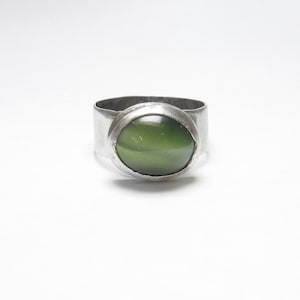 May include: A silver ring featuring a wide band and a large, oval, olive-green stone. The stone is set in a bezel setting, and the ring has a simple, minimalist design. The ring is likely made of sterling silver.