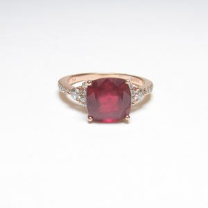 May include: A rose gold ring featuring a large, cushion-cut deep red ruby as the center stone. The band is accented with small, round, clear white diamonds set along the shoulders, leading up to the central ruby held by four metal prongs.