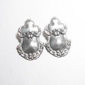May include: A pair of silver-coloured earrings with an ornate design. Each earring has a shield-like form with floral and scrollwork details. The design includes a central, slightly convex area and a decorative top.