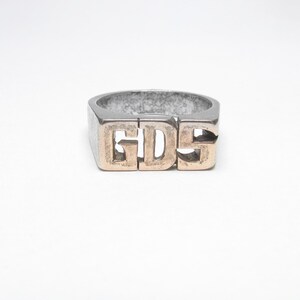 May include: A silver-toned ring with the letters "GDS" in a contrasting gold color. The ring has a rectangular shape and a slightly worn appearance, set against a white background.