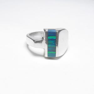 May include: A silver ring featuring a smooth, polished metal band and a wide, flat face. A vertical rectangular inlay of iridescent blue and green opal-like material is set into the center of the ring face, contrasting with the metallic surface.