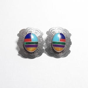 May include: Pair of silver-toned earrings with an oval, multi-colored inlay. The inlay features sections of blue, turquoise, red, green, purple, and yellow. The earrings have a sunburst design around the oval inlay.
