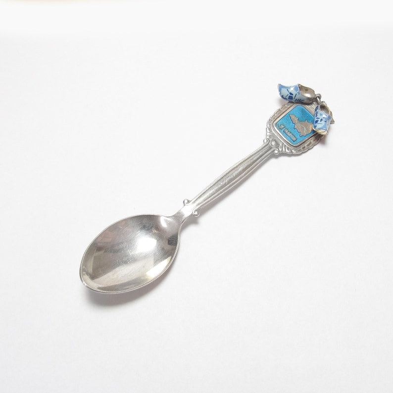 Pode incluir: Colher de souvenir de prata com um design de esmalte azul e branco de um mapa dos Pa&iacute;ses Baixos e dois tamancos azuis e brancos. O cabo da colher &eacute; decorado com um design floral. A colher diz "Holanda" no cabo.