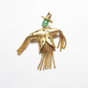 Op de afbeelding: Gouden  knuppel  broche met een groen stenen gezicht en gouden franjes.