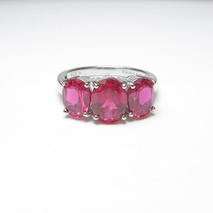 May include: A silver ring featuring three oval-cut, vibrant pink stones set in a prong setting. The inner band is clearly stamped with the text 925 and THAILAND, indicating the metal composition and origin of this piece of jewelry.