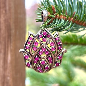 May include: A silver pendant in the shape of a lotus flower, adorned with numerous small, round, pink gemstones. The pendant is suspended from a silver bail and is set against a backdrop of green pine needles.