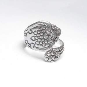 May include: A silver spoon ring with detailed floral and grape cluster patterns. The ring has an adjustable wrap-around design, displaying the ornate designs of the repurposed silverware. The ring is set against a white background.