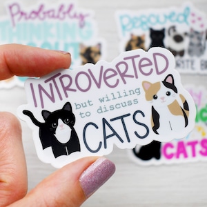 May include: A white sticker with black and purple text that reads "Introverted but willing to discuss cats". The sticker features two cartoon cats, one black and white and one orange and white.