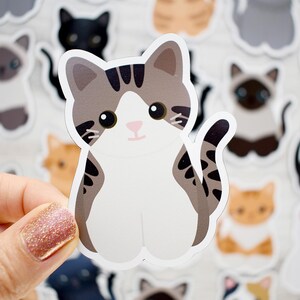 May include: A die-cut vinyl sticker featuring a cartoon-style cat with grey and brown tabby markings on its head and tail, a white face, chest, and paws, and large black eyes. The cat is depicted in a sitting position with its tail curled upward.