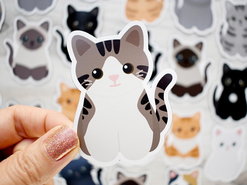 May include: A die-cut vinyl sticker featuring a cartoon-style cat with grey and brown tabby markings on its head and tail, a white face, chest, and paws, and large black eyes. The cat is depicted in a sitting position with its tail curled upward.