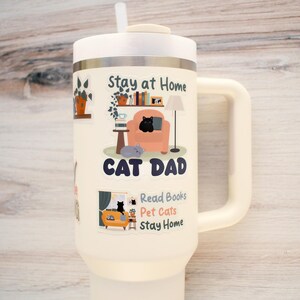 K&ouml;nnte beinhalten: Cremefarbener Isolierbecher mit wei&szlig;em Deckel und Griff. Der Becher zeigt Illustrationen zum Thema Katze und den Text "Stay at Home, Cat Dad, Read Books, Pet Cats, Stay Home."