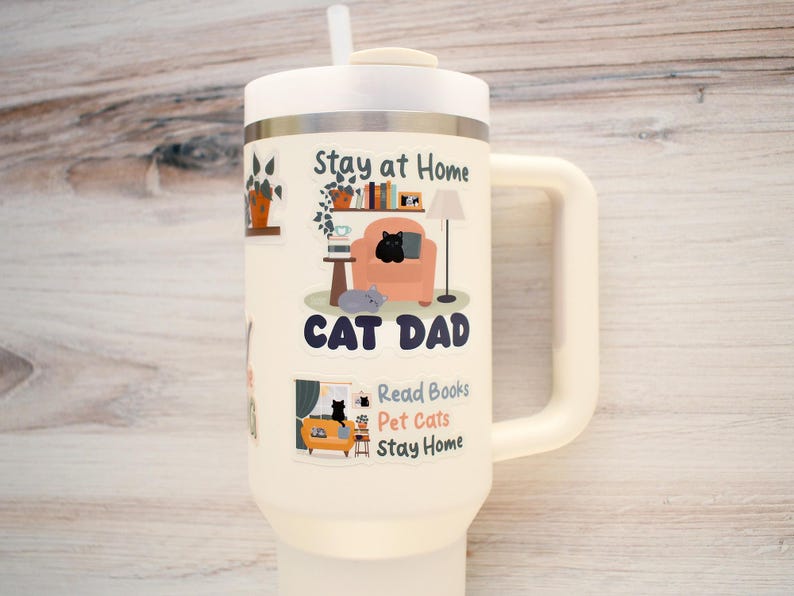 K&ouml;nnte beinhalten: Cremefarbener Isolierbecher mit wei&szlig;em Deckel und Griff. Der Becher zeigt Illustrationen zum Thema Katze und den Text "Stay at Home, Cat Dad, Read Books, Pet Cats, Stay Home."