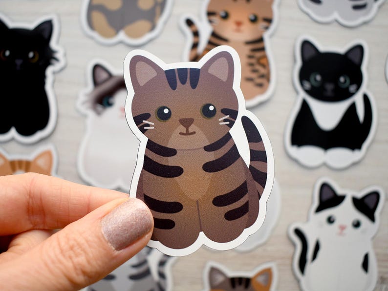 May include: A die-cut vinyl sticker featuring a cartoon brown tabby cat with dark brown stripes, large black eyes, and a small nose. The cat is sitting in a forward-facing position with its tail curled to the side, surrounded by other cat stickers.