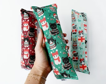 May include: Three cat toys with Christmas-themed patterns. The toys are rectangular and filled with catnip. One is green with white cats in sweaters, another is red with white cats, and the third is light blue with gray cats.