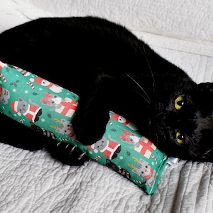 May include: A black cat is lying on a white surface, cuddling a green cat toy. The toy is rectangular and covered in a festive pattern of cats, gifts, and holiday imagery.