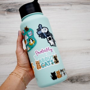 May include: A light blue reusable water bottle with various cat-themed stickers, including a sticker that says "Probably Thinking About Cats".