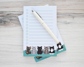 May include: A notepad with a teal border featuring cartoon cats. A cream-colored pen rests on the notepad. The notepad has a checklist and lined paper. The background is a light wood grain.