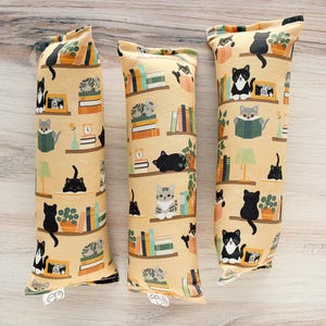 May include: Three catnip toys with a repeating pattern of cats, bookshelves, and lamps on a beige background. The toys are cylindrical and filled with catnip, designed for feline entertainment.