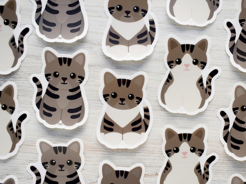 May include: A collection of die-cut stickers featuring cartoon-style tabby cats in various poses. Each sticker displays a cat with brown and gray striped fur, large dark eyes, and a small pink nose, arranged in a repeating pattern on a white surface.