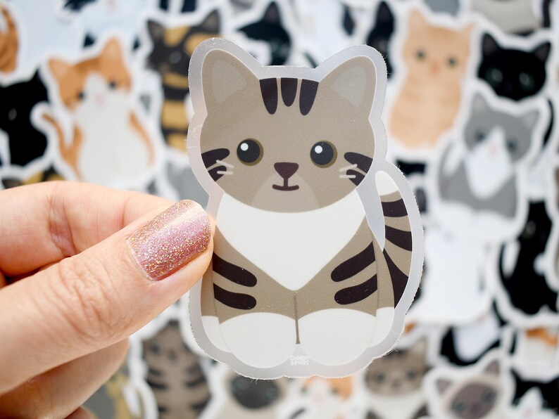May include: A die-cut vinyl sticker featuring a cartoon-style brown and white tabby cat with large black eyes, dark brown stripes on its head and body, and white paws. The cat is sitting in an upright position with its tail curled to the side.