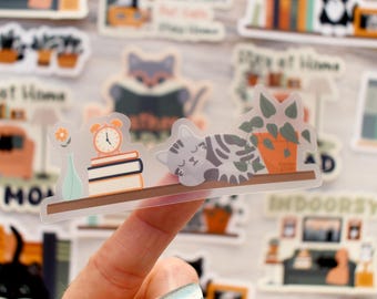 Könnte beinhalten: Ein klarer Aufkleber mit einer schlafenden grauen Tabby-Katze auf einem Regal mit einer Topfpflanze, Büchern und einem Wecker. Der Aufkleber hat ein gemütliches, häusliches Thema mit einer hellen Farbpalette. Andere Aufkleber sind im Hintergrund sichtbar.