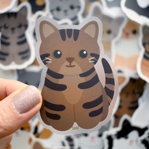 May include: A die-cut vinyl sticker featuring a brown tabby cat with dark brown stripes, large black eyes, and a small nose. The cat is depicted in a seated position with its tail curled to the side. The text Clover & Pearl is printed in small letters at the bottom.