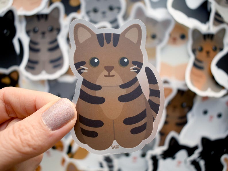 May include: A die-cut vinyl sticker featuring a brown tabby cat with dark brown stripes, large black eyes, and a small nose. The cat is depicted in a seated position with its tail curled to the side. The text Clover & Pearl is printed in small letters at the bottom.