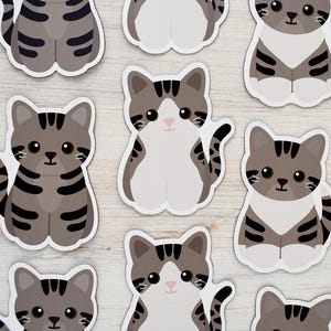 May include: A collection of die-cut stickers featuring cartoon-style gray tabby cats with dark stripes, white chests, and large black eyes. The cats are depicted in a seated position with their tails curled, showcasing a repeating pattern of feline designs.
