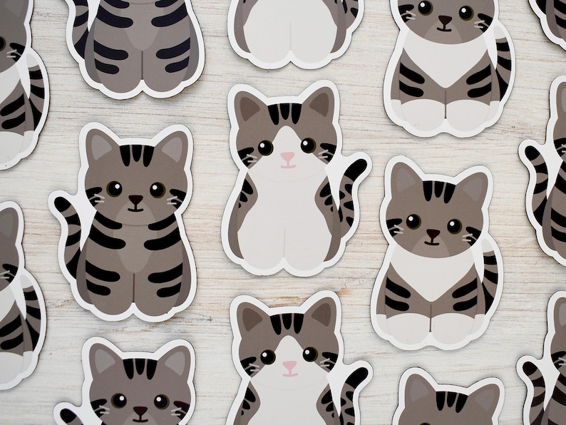 May include: A collection of die-cut stickers featuring cartoon-style gray tabby cats with dark stripes, white chests, and large black eyes. The cats are depicted in a seated position with their tails curled, showcasing a repeating pattern of feline designs.