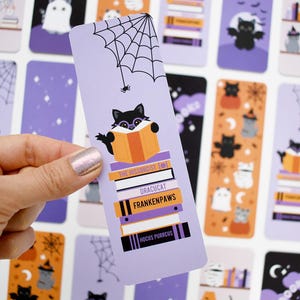 May include: A Halloween-themed bookmark featuring a black cat wearing glasses and reading a book. The bookmark has a spiderweb and spider design at the top, and the book titles include "Dracucat" and "Frankenpaws."