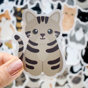 May include: A die-cut sticker featuring a cartoon brown tabby cat with dark brown stripes, large black eyes, and a small nose. The cat is sitting in a forward-facing position with its tail curved upward. The sticker has a white border outline.