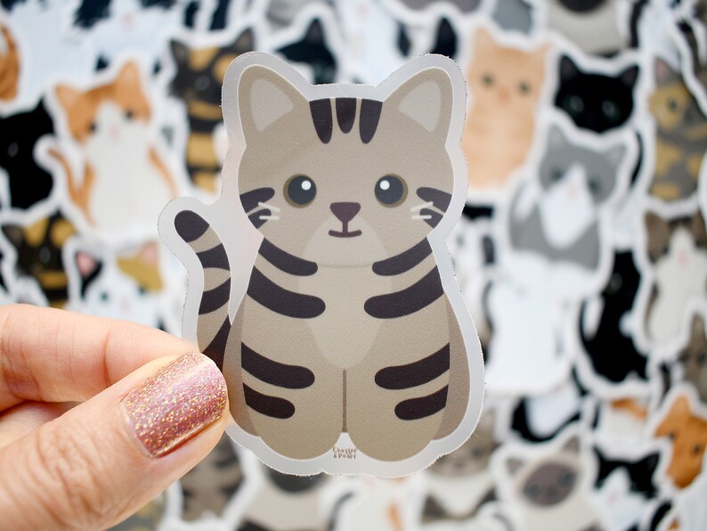 May include: A die-cut sticker featuring a cartoon brown tabby cat with dark brown stripes, large black eyes, and a small nose. The cat is sitting in a forward-facing position with its tail curved upward. The sticker has a white border outline.