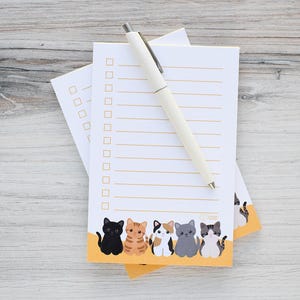 May include: A stack of white notepads with a row of cartoon cats at the bottom. A cream-colored pen rests on top of the notepads. The notepads have a checklist design with horizontal lines and check boxes.