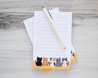 May include: A stack of white notepads with a row of cartoon cats at the bottom. A cream-colored pen rests on top of the notepads. The notepads have a checklist design with horizontal lines and check boxes.