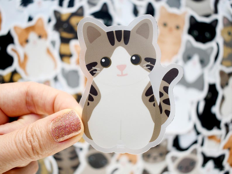 May include: A die-cut sticker featuring a cartoon cat with brown tabby markings on its head and tail, a white face, chest, and belly, and large black eyes. The cat is shown in a sitting position with its tail curled to the side against a background.