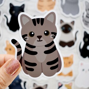 May include: A die-cut sticker featuring a cartoon gray tabby cat with dark gray stripes, large black eyes, and a small nose. The cat is in a sitting position with its tail curved upward. The sticker has a white border and a matte finish.
