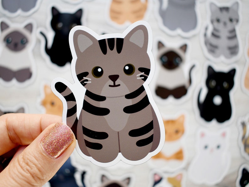 May include: A die-cut sticker featuring a cartoon gray tabby cat with dark gray stripes, large black eyes, and a small nose. The cat is in a sitting position with its tail curved upward. The sticker has a white border and a matte finish.