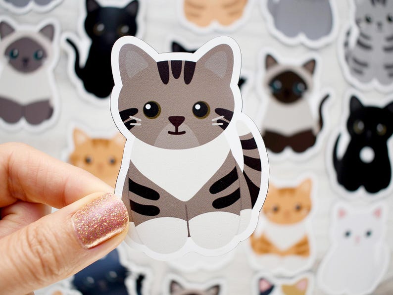 May include: A die-cut sticker featuring a cartoon brown and gray tabby cat with dark stripes, large black eyes, and white paws. The cat is sitting in a forward-facing position with a white chest patch and a striped tail curled to the side.
