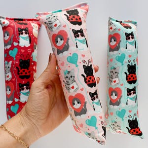 May include: Three catnip toys with colorful patterns. One is held by a hand. The toys feature repeating patterns of cartoon cats, hearts, and ladybugs on red, pink, and teal backgrounds. The cats wear sunglasses and heart-shaped collars.