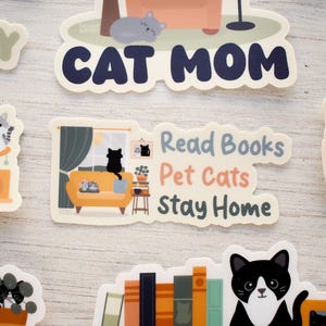 K&ouml;nnte beinhalten: Eine Sammlung von Aufklebern zum Thema Katze mit verschiedenen Designs. Ein Aufkleber zeigt "Cat Mom" in Marineblau. Ein anderer zeigt eine Katze auf einem gelben Sofa mit dem Text "Read Books, Pet Cats, Stay Home". Weitere zeigen Katzen und B&uuml;cher.