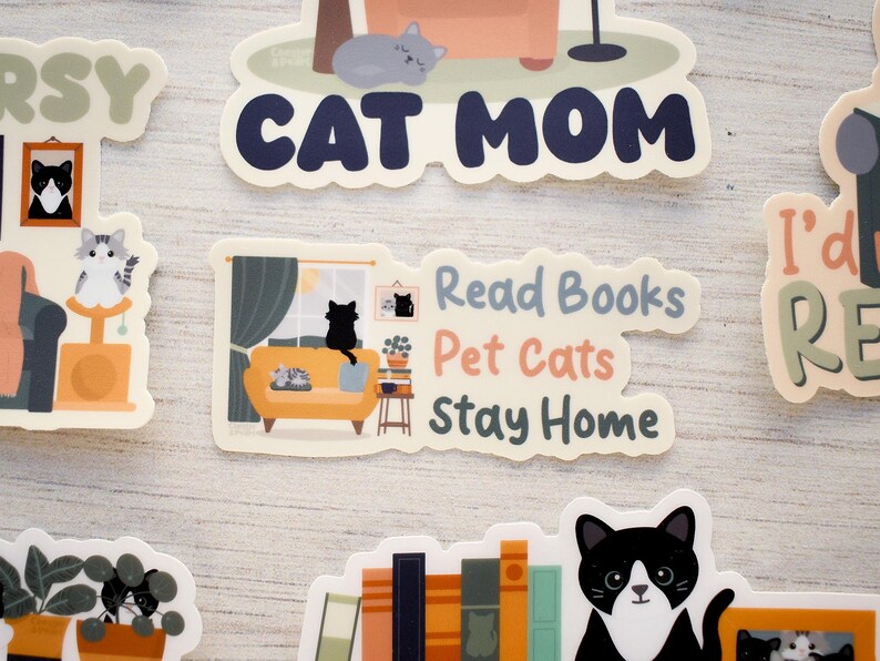 K&ouml;nnte beinhalten: Eine Sammlung von Aufklebern zum Thema Katze mit verschiedenen Designs. Ein Aufkleber zeigt "Cat Mom" in Marineblau. Ein anderer zeigt eine Katze auf einem gelben Sofa mit dem Text "Read Books, Pet Cats, Stay Home". Weitere zeigen Katzen und B&uuml;cher.