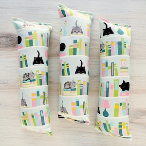 May include: Three catnip toys with a light blue background, featuring stacked book designs and various cat illustrations. The toys have a cylindrical shape and are decorated with colorful books, lamps, and cats in different poses.