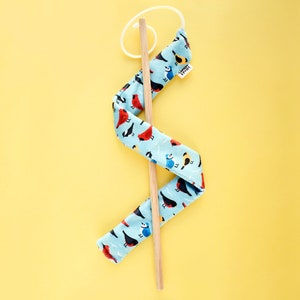 May include: A blue fabric cat toy with a bird pattern, attached to a wooden stick with a white elastic loop. The fabric features red, black, and blue birds on a light blue background. A small tag is attached to the fabric.