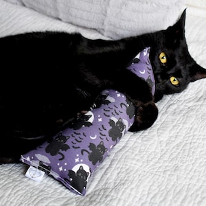 May include: A black cat with yellow eyes plays with a purple catnip toy. The toy is patterned with black bats, moons, and stars. The cat is lying on a white quilted surface.