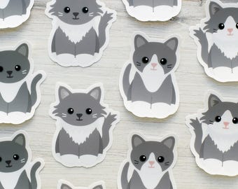 May include: A collection of die-cut stickers featuring a cartoon cat with grey and white fur, large black eyes, and a small pink nose. Each sticker depicts the cat in a seated position with a white chest, white paws, and a grey tail.