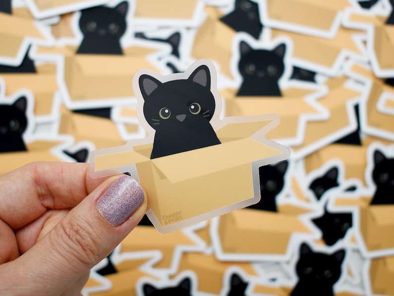 May include: A black cat peeking out of a brown cardboard box. The cat is looking directly at the viewer. The sticker is clear and has a white border.