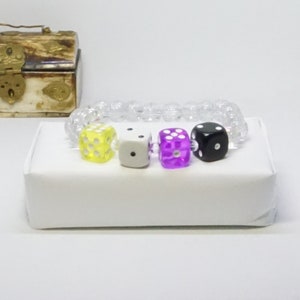 May include: A clear beaded bracelet with four dice charms. The dice are yellow, white, purple, and black.